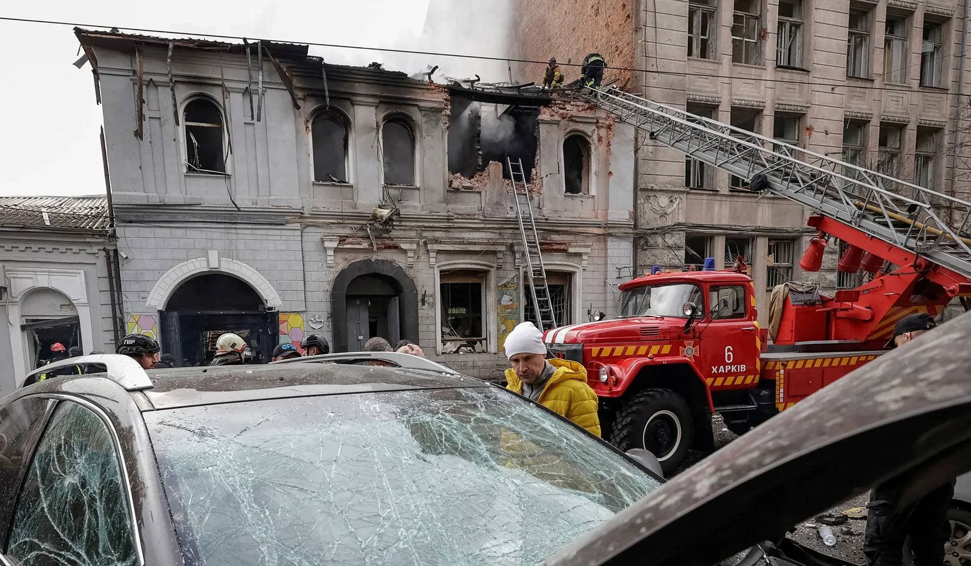 A resident looks at a damaged car as firefighters work at the site of the kindergarten hit by Russian drone strike in Kharkiv